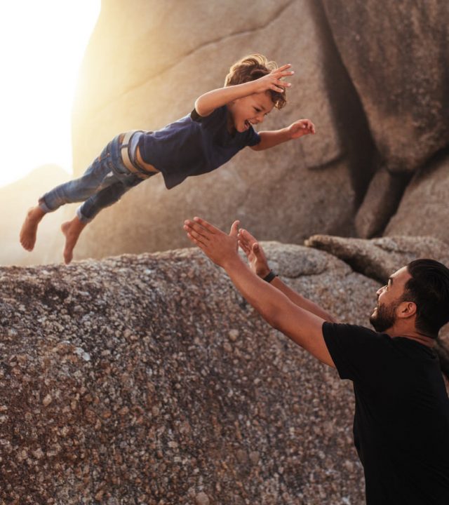 Shot of cute little boy leaping into his fathers arms from a big rock. Father and son enjoying summer holidays at the beach.